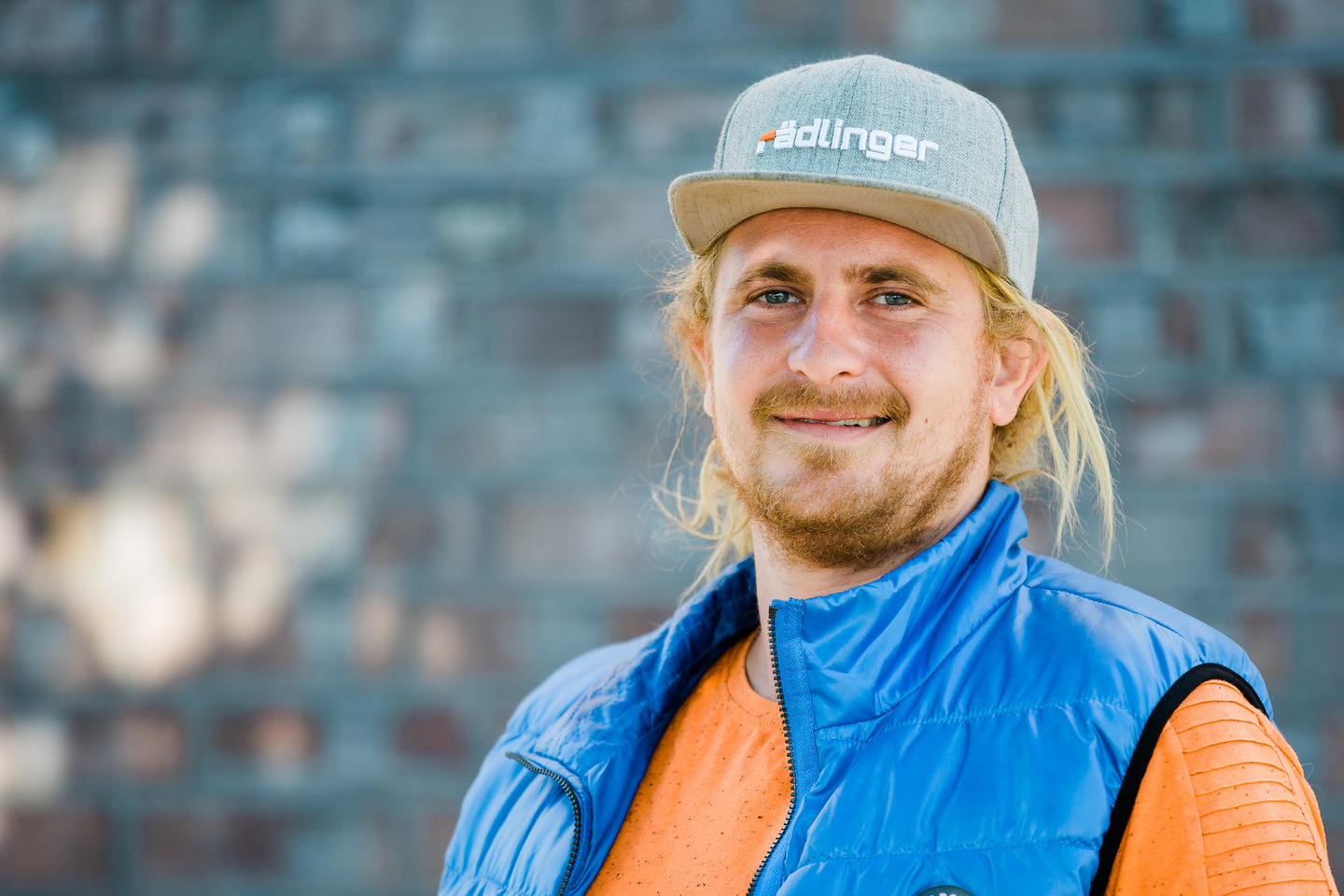 Ein Mann mit blonden Haaren, Bart und Lächeln trägt eine graue Kappe mit dem Logo "Adlinger" und eine blaue Weste über einem orangefarbenen Oberteil.
